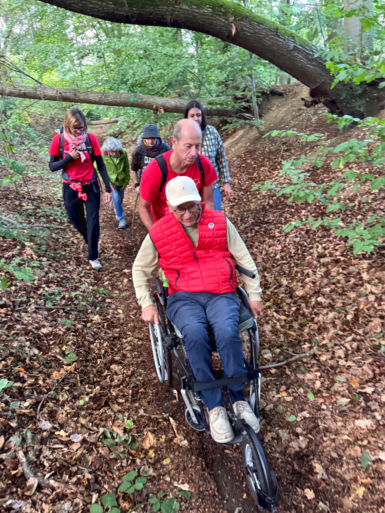 Un groupe de randonneurs et de bénévoles marchent dans chemin au milieu d'un bois avec une personne en fauteuil roulant poussée par un bénévole